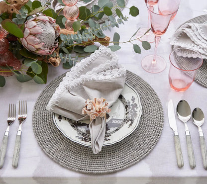 The Shell Edge napkin in gray with hand-crocheted raffia edging in white. A versatile napkin suited for bohemian and rustic-inspired table settings. 100% linen with raffia trimming.