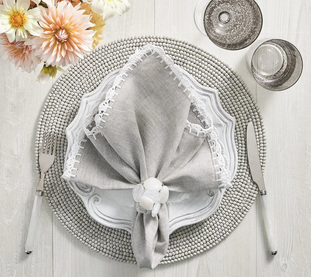 The Shell Edge napkin in gray with hand-crocheted raffia edging in white. A versatile napkin suited for bohemian and rustic-inspired table settings. 100% linen with raffia trimming.